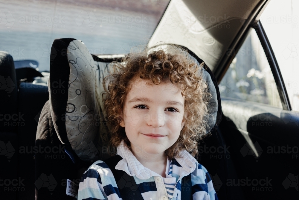 Happy child strapped into the child seat of the family car outside home - Australian Stock Image