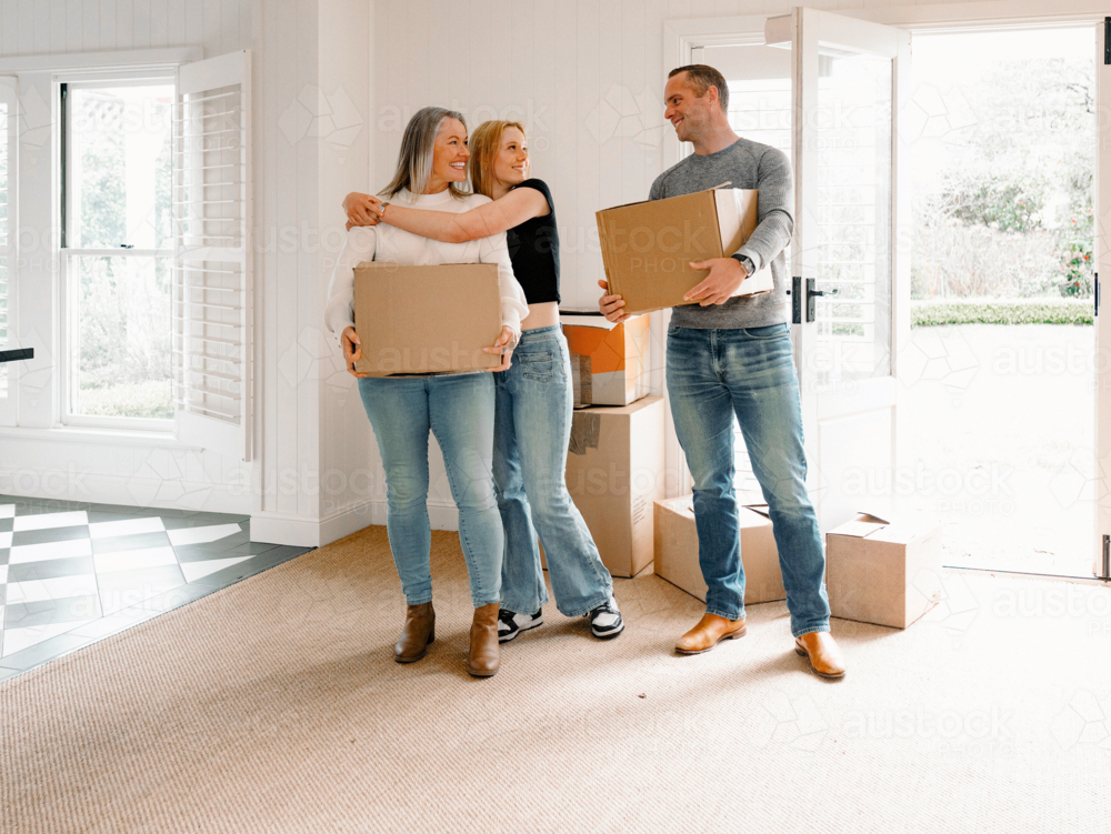 Happy couple carrying cardboard boxes while teenage daughter hugs mum from behind inside home - Australian Stock Image
