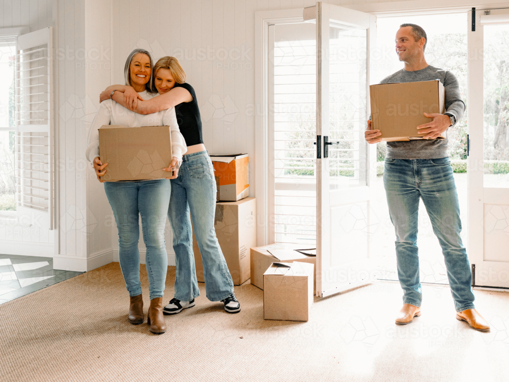 Happy couple carrying cardboard boxes while teenage girl hugs mum from behind inside home - Australian Stock Image