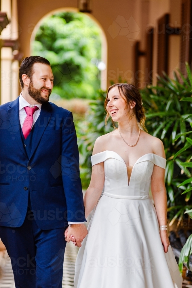 Happy couple laughing and smiling at one another as they hold hands on their wedding day - Australian Stock Image