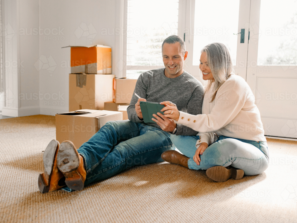 Happy couple seated on carpeted floor of their new home holding a tablet device - Australian Stock Image