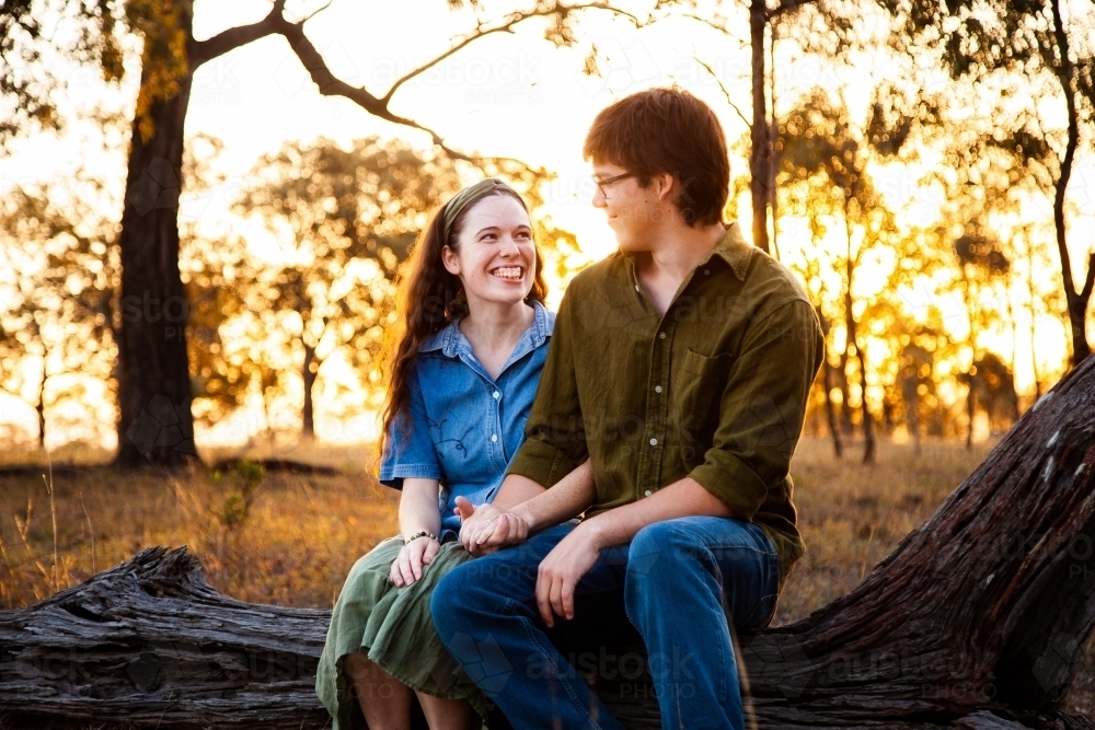 Happy couple sitting on a log in paddock at sunset - Australian Stock Image