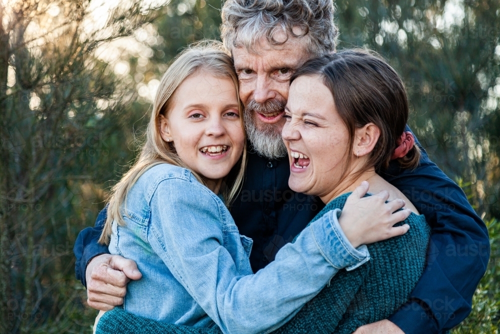 Happy dad with his two teenage daughters hug together outside - Australian Stock Image