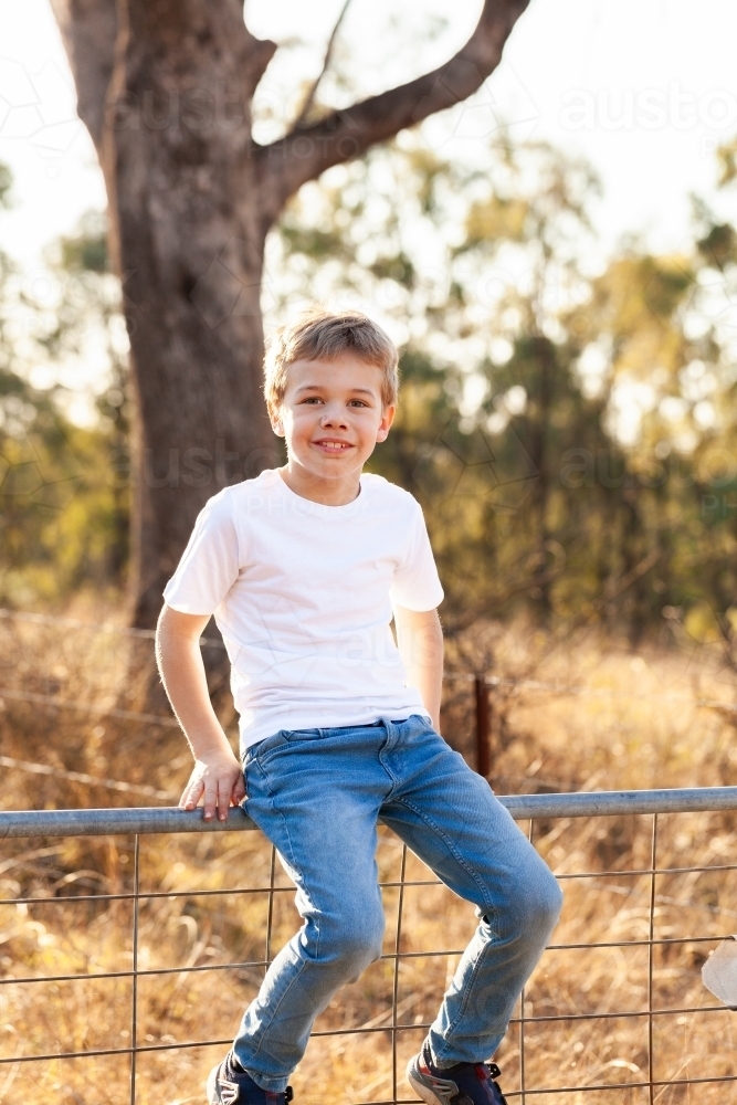 Happy eight year old Australian kid sitting on gate in countryside - Australian Stock Image