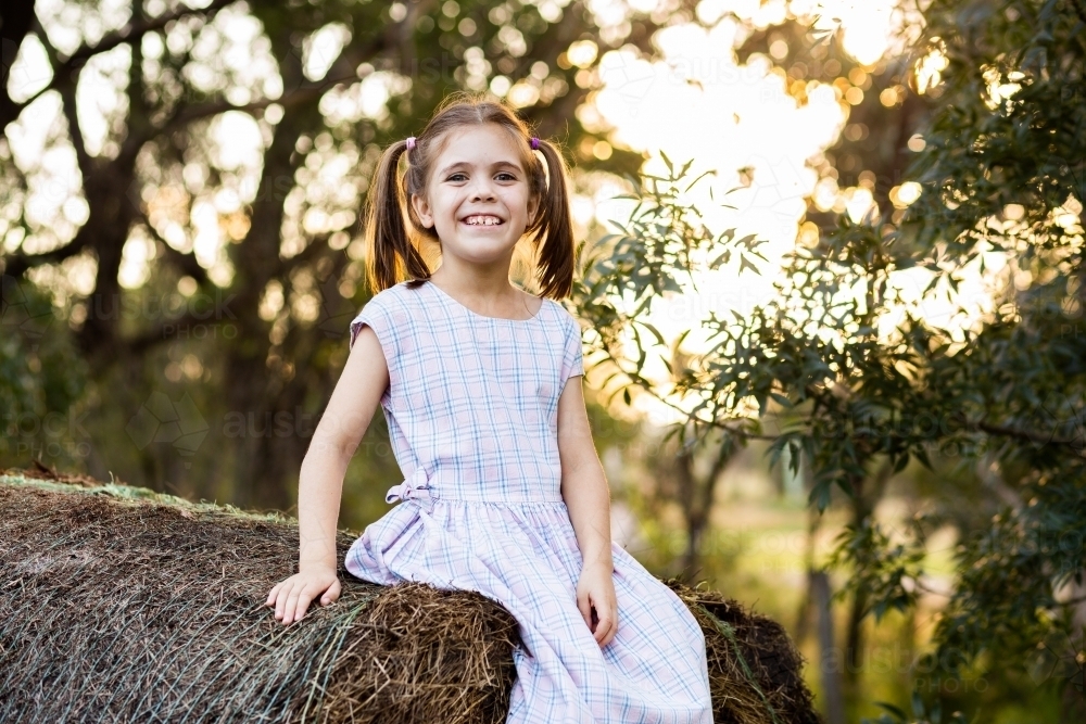 Happy eight year old country girl sitting on round hay bale - Australian Stock Image