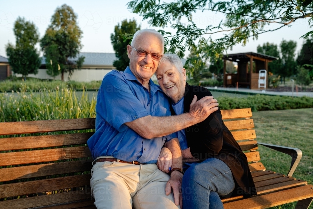 Happy, elderly retired married couple hugging on a park bench - Australian Stock Image