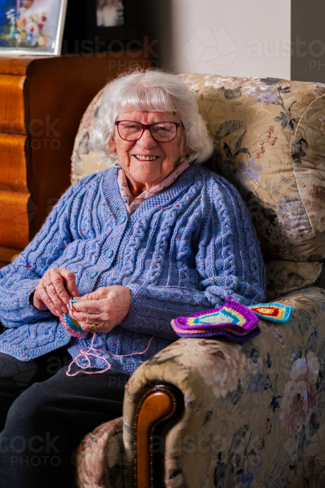 happy elderly woman sitting on armchair crocheting blanket square for great grandchild - Australian Stock Image