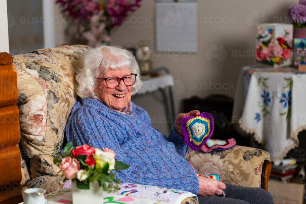happy elderly woman sitting on armchair crocheting blanket square for great grandchild - Australian Stock Image