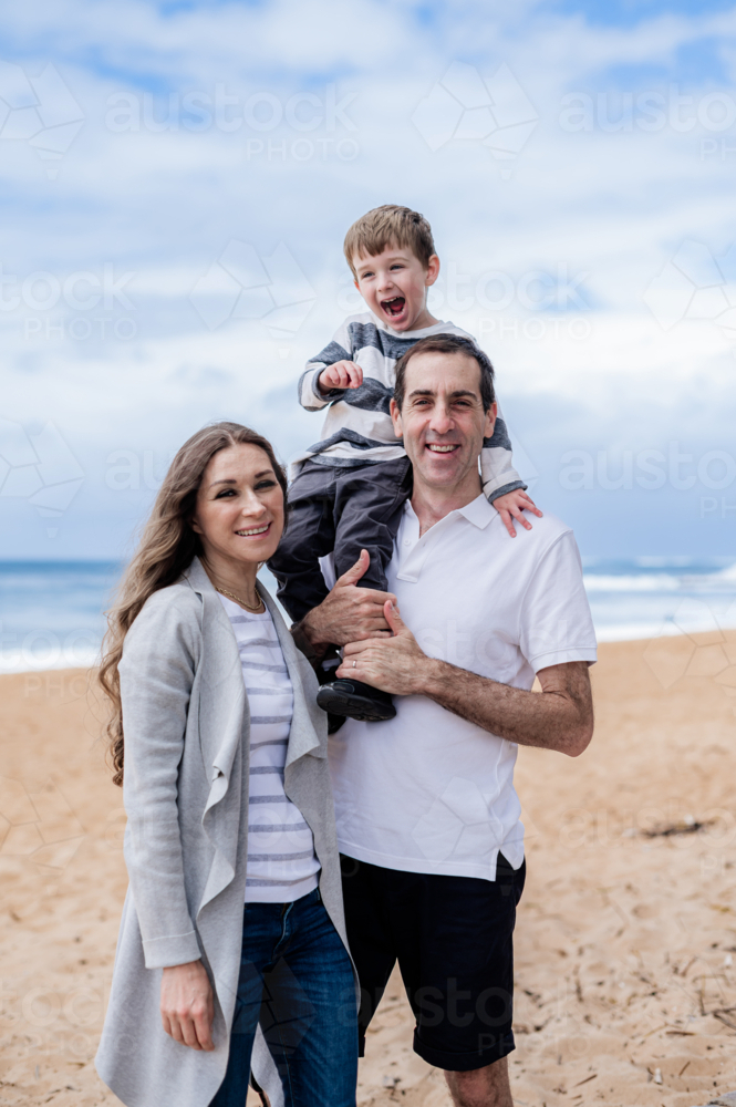 Happy family enjoys a sunny day at the beach, with a boy gleefully on his father's shoulders - Australian Stock Image