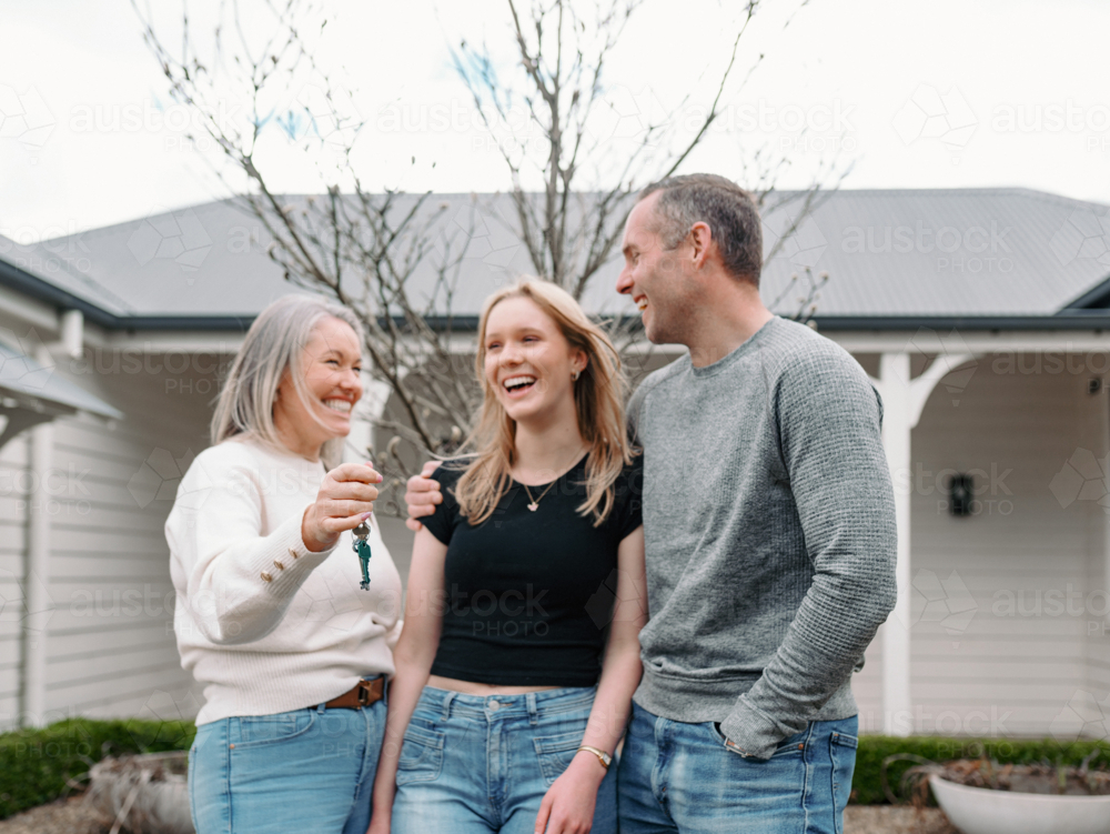 Happy family standing in front yard with mum holding out the key of their new home - Australian Stock Image