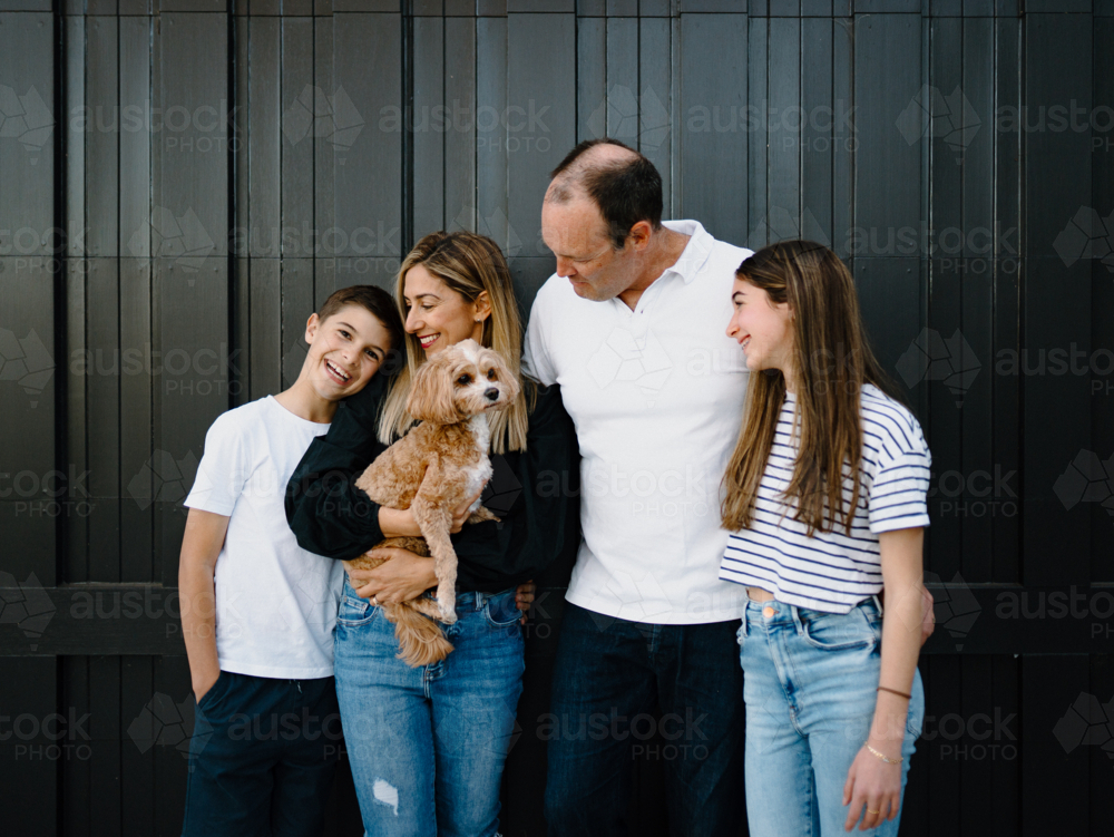 Happy family standing outside in front of black garage door with pet dog - Australian Stock Image