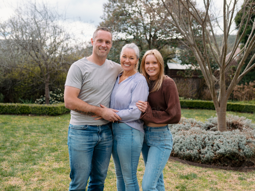Happy family stands closely hugging each other in the garden of their new home - Australian Stock Image