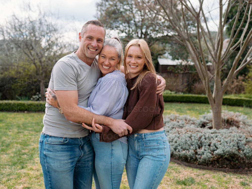 Happy family stands closely hugging each other in the garden of their new home - Australian Stock Image