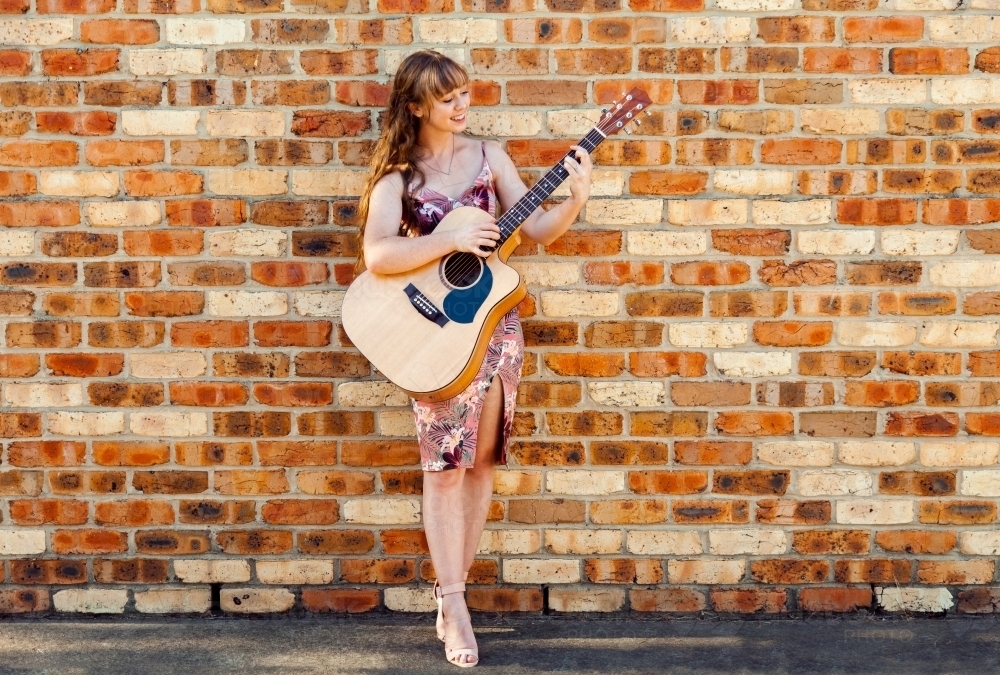 Happy female musician with guitar against brick wall - Australian Stock Image