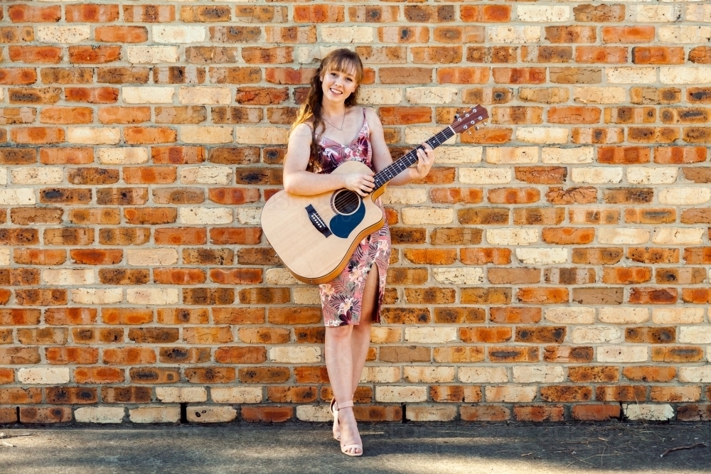 Happy female musician with guitar against brick wall - Australian Stock Image