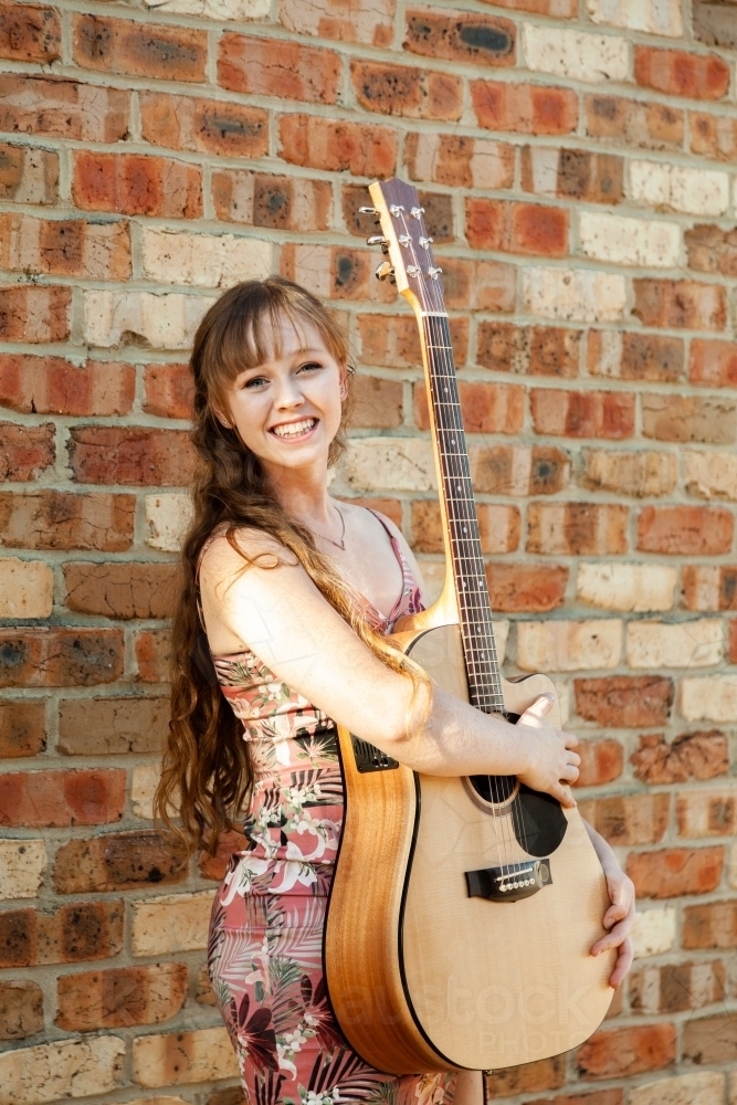 Happy female musician with guitar against brick wall - Australian Stock Image