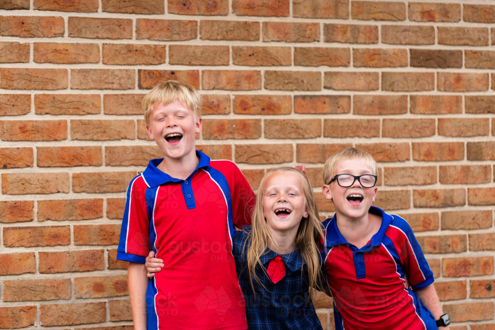 Happy group of three primary school kids laughing together with arms around each other - Australian Stock Image