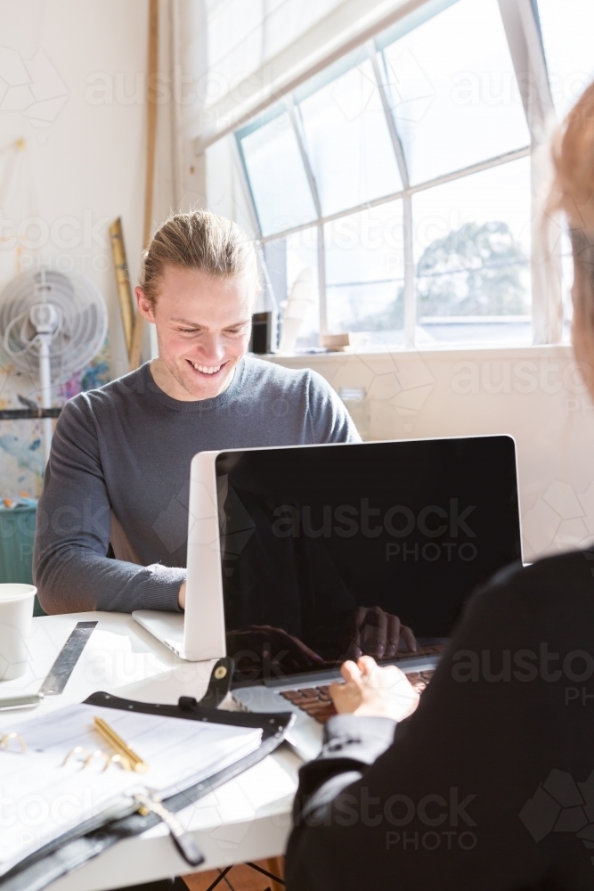 Happy guy working on his laptop in a studio opposite a colleague - Australian Stock Image