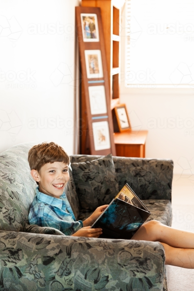 Happy kid reading a book on lounge at home - Australian Stock Image