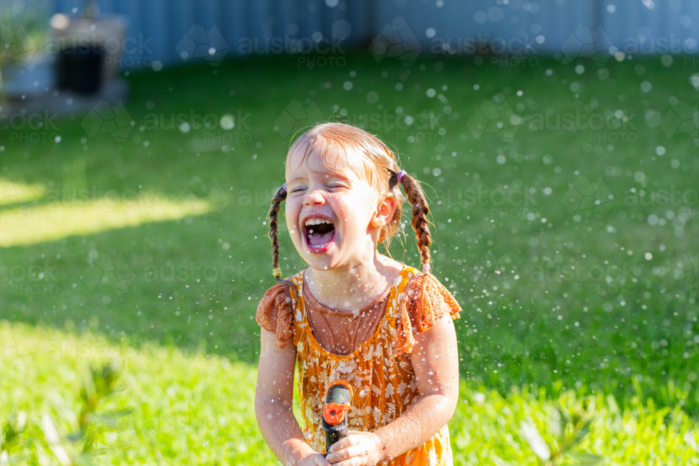 Happy laughing little girl joyfully playing with sprinkler hose watering herself in garden - Australian Stock Image