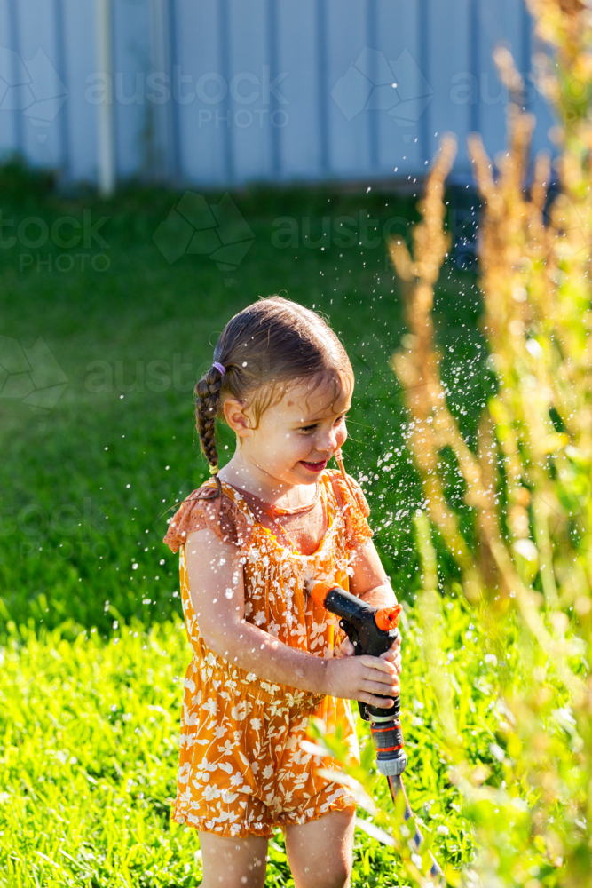 Happy laughing little girl playing with sprinkler hose watering herself in garden - Australian Stock Image