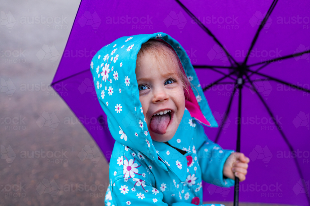 Happy little Aussie girl on rainy day wearing raincoat holding purple umbrella with her tongue out - Australian Stock Image