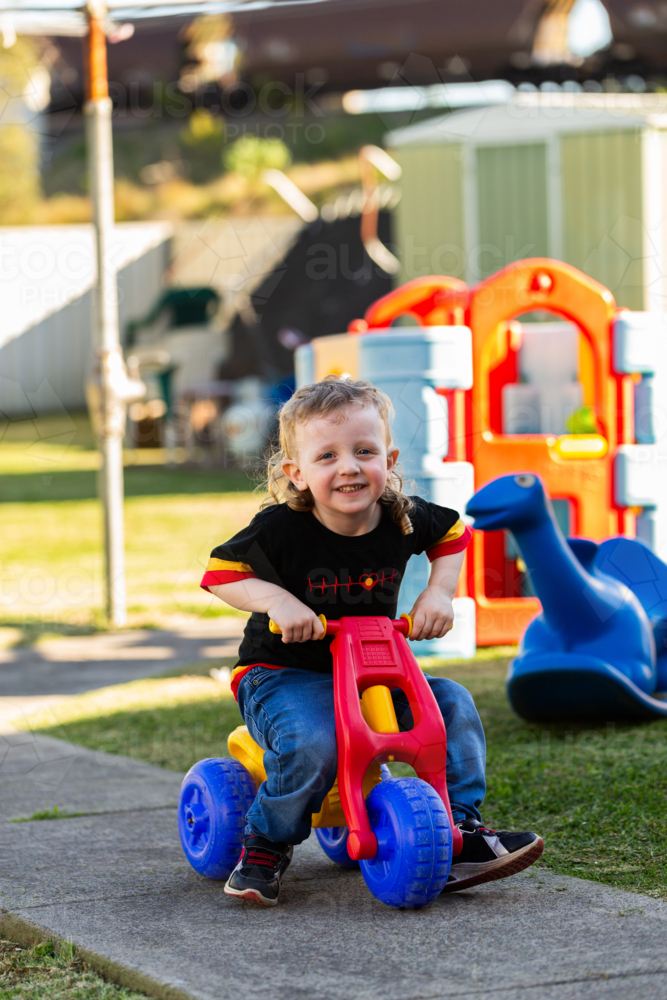 Happy little Aussie kid playing with colourful trike in backyard of home - Australian Stock Image