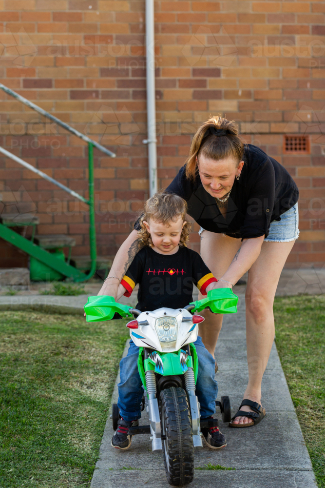 Happy little Aussie kid riding on motorbike balance bike in backyard of home with mum helping - Australian Stock Image