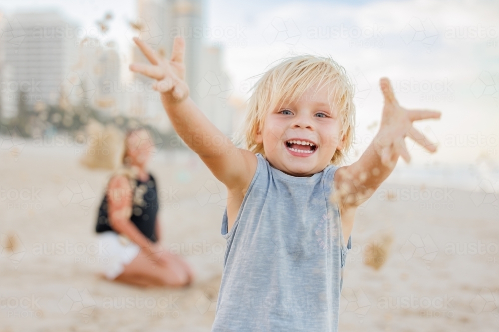 Happy little boy throwing sand on the beach with mother in background - Australian Stock Image