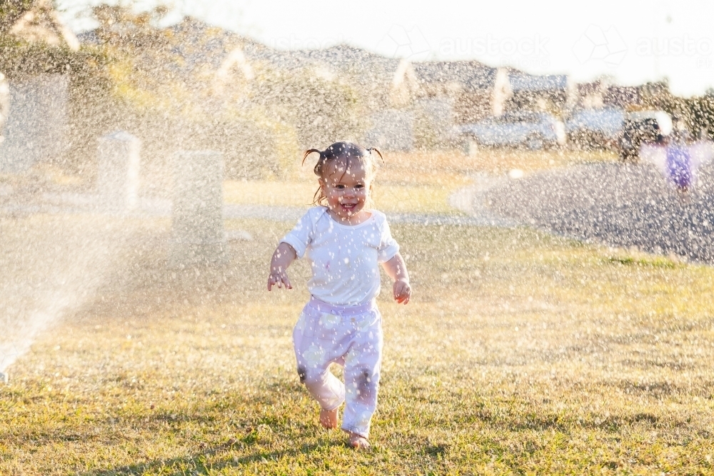 Happy little girl getting wet under sprinkler in front yard of suburban home - Australian Stock Image