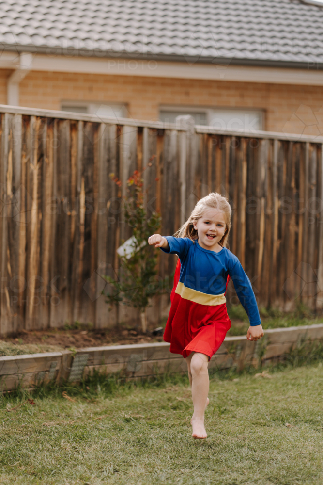 Happy little girl in a superhero costume running around their yard. - Australian Stock Image