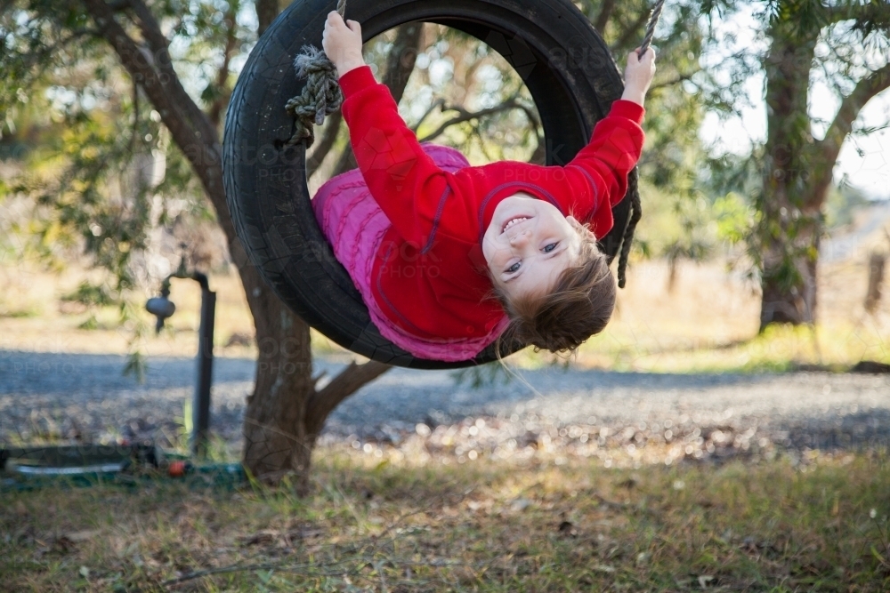Happy little girl on a tire swing looking at the camera in the backyard - Australian Stock Image
