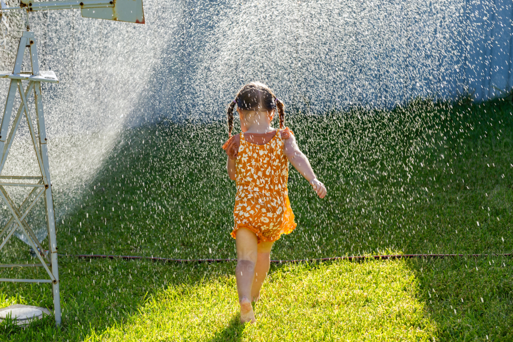 Happy little girl playing under sprinkler hose in garden of suburban backyard - Australian Stock Image