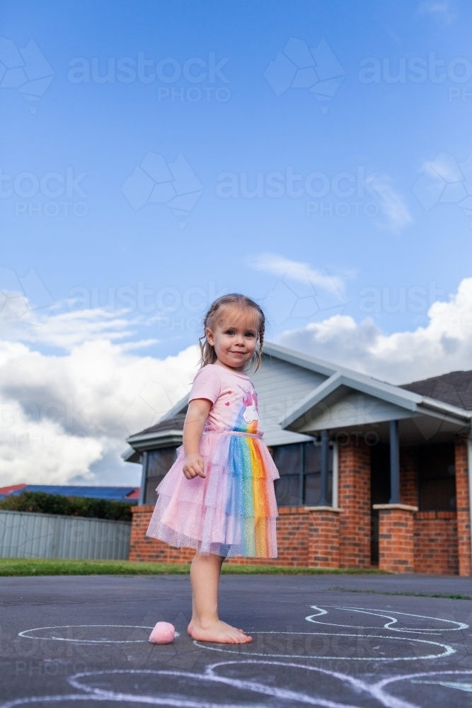 Happy little girl sanding on suburban driveway with chalk   - Australian Stock Image