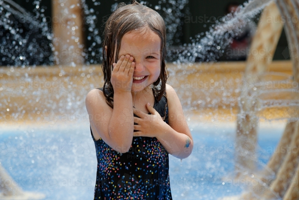 Happy little girl under fountains at a water park, wiping her eye - Australian Stock Image