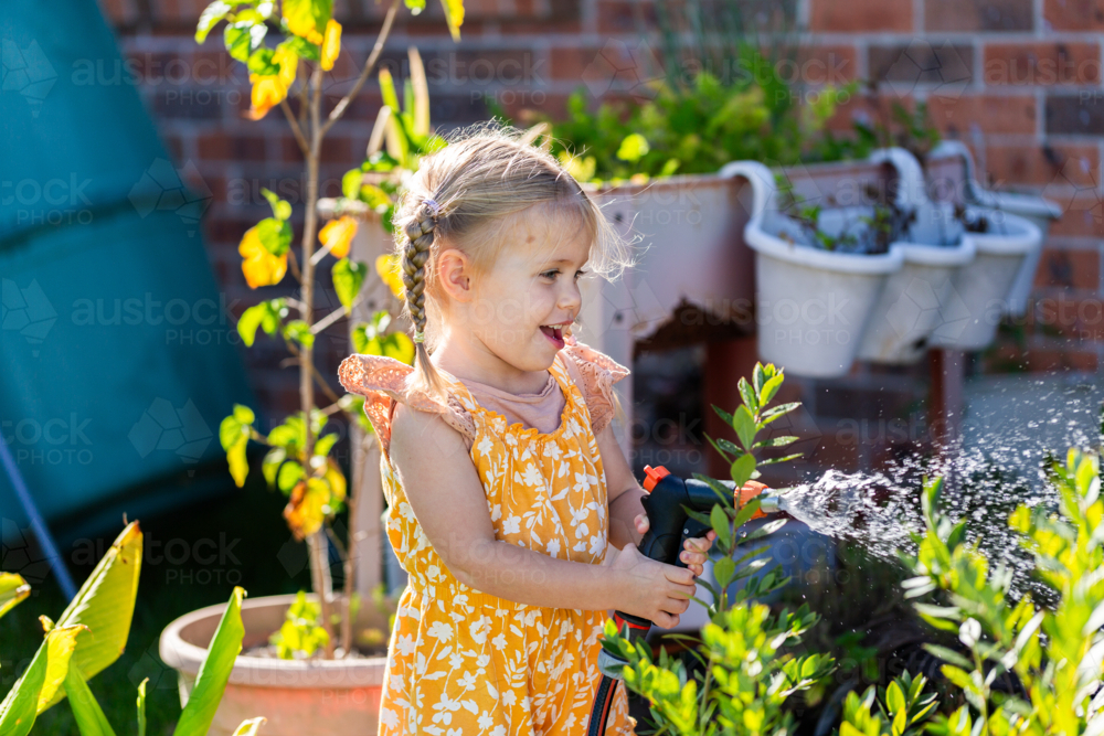 Happy little girl watering plants in garden at home with spray nozzle on hose - Australian Stock Image
