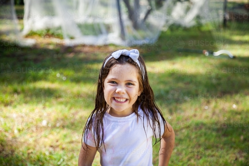 Happy little girl wet and grinning after running under garden sprinkler - Australian Stock Image