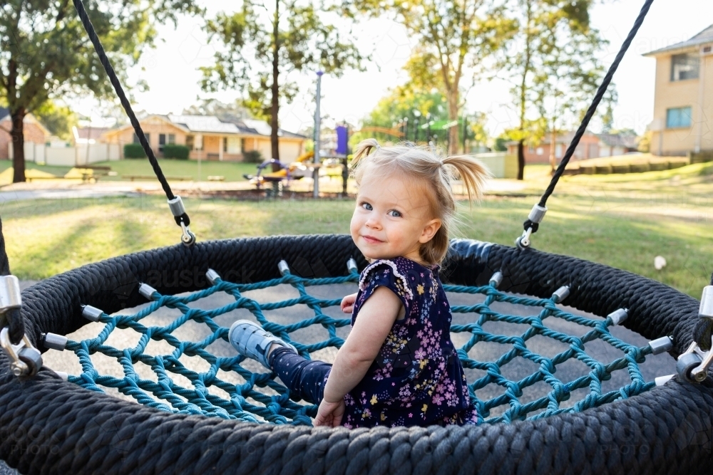 Happy little toddler girl looking back over shoulder on swing at park - Australian Stock Image