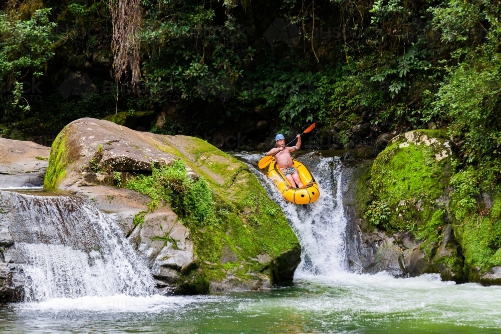 Happy man taking his inflatable kayak down a small waterfall in a mountain river - Australian Stock Image