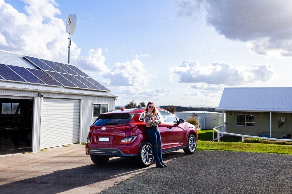 happy middle age woman standing with electric vehicle car and solar panels - sustainable living - Australian Stock Image