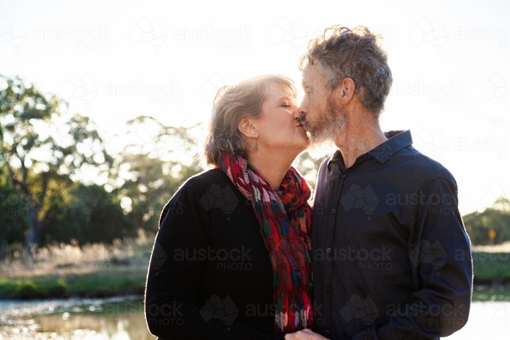 Happy middle aged couple kiss beside dam on farm - Australian Stock Image
