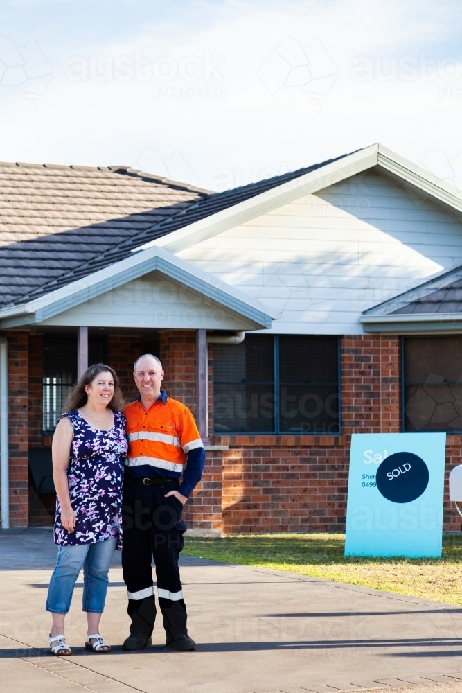 Happy middle aged couple standing infront of house with sold real estate sign - Australian Stock Image