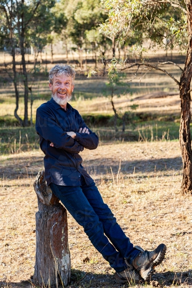Happy middle aged man with beard leaning on stump in paddock - Australian Stock Image