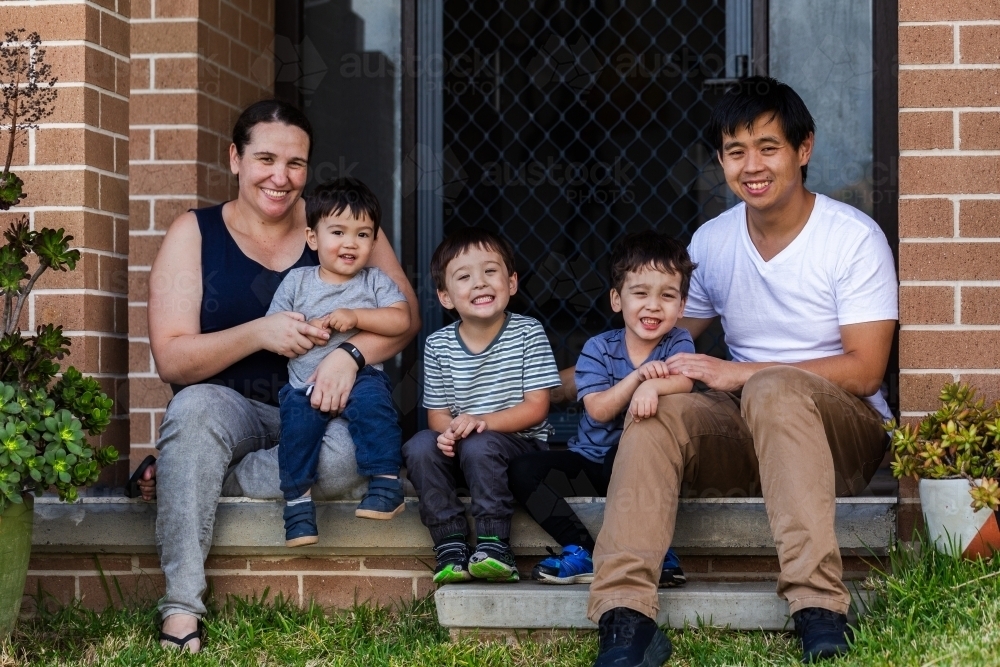 Happy mixed race Chinese Australian family sitting together on front doorstep of home - Australian Stock Image