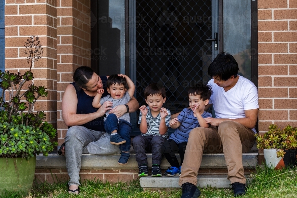 Happy mixed race Chinese Australian family sitting together on front doorstep of home - Australian Stock Image