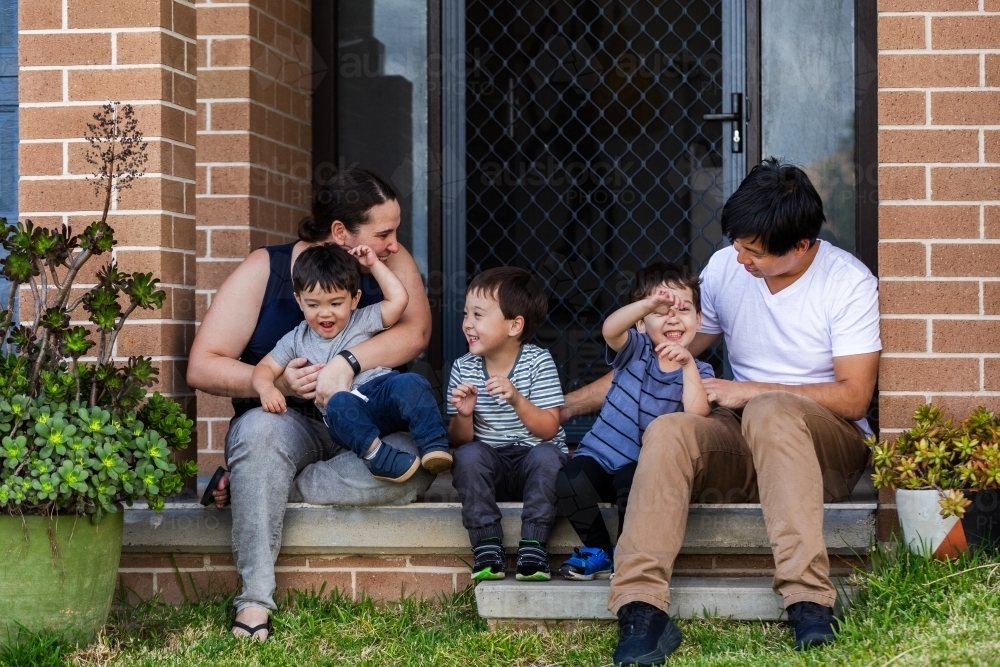 Happy mixed race Chinese Australian family sitting together on front doorstep of home - Australian Stock Image