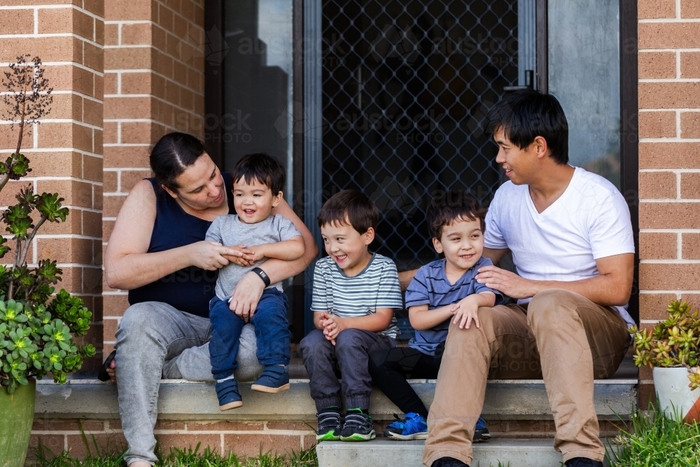 Happy mixed race Chinese Australian family sitting together on front doorstep of home - Australian Stock Image