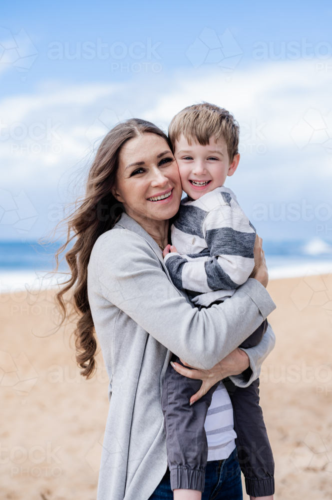 Happy mother holds her smiling son at the beach, both enjoying a bright and sunny day outdoors - Australian Stock Image