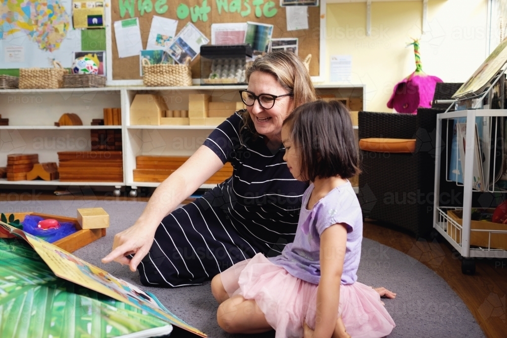 Happy multicultural teacher and child reading a book in kindergarten - Australian Stock Image