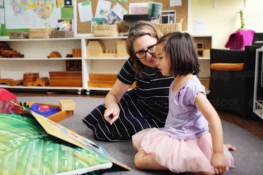 Happy multicultural teacher and child reading a book in kindergarten - Australian Stock Image